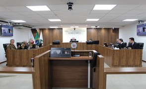 A imagem mostra os sete desembargadores do TJMMG na sala do Tribunal Pleno. Quatro vestem farda militar, e três vestem toga. Eles estão sentados atrás de mesas de madeira com computadores em cima. Em primeiro plano há uma tribuna madeira com uma tela, e ao fundo há um letreiro com o nome por extenso do TJMMG