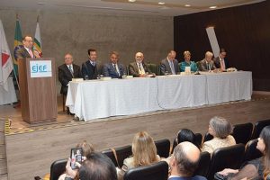 A imagem mostra 7 homens e uma mulher em lugar de destaque no lançamento do livro “200 anos do Direito Constitucional Brasileiro”. Eles estão sentados atrás de uma mesa comprida, ouvindo o discurso do presidente do TJMG, desembargador Luiz Carlos Corrêa Junior, que veste terno marrom, gravata vermelha e fala ao microfone atrás de um púlpito de madeira. A segunda pessoa em destaque é o ouvidor do TJMMG, desembargador Fernando José Armando Ribeiro usando terno preto, gravata roxa e suéter marrom. A parede ao fundo é cinza
