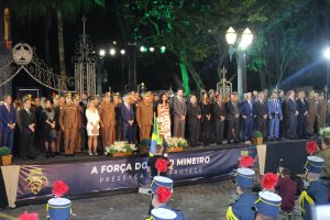 A imagem mostra o desembargador James Ferreira Santos na primeira fileira de autoridades em cima de um palanque. Ele veste terno cinza e gravata vermelha. Na segunda fileira está o desembargador Rúbio Paulino Coelho usando terno cinza e gravata vermelha. Ao redor deles há diversas autoridades entre homens e mulheres que vestem farda militar oficial e roupas civis formais. Em primeiro plano há representantes da PMMG. À frente do palanque há uma faixa escrito “A força do povo mineiro, presença que protege”
