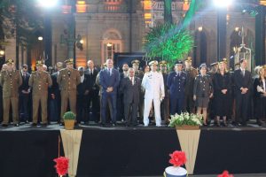 A imagem mostra o presidente do TJMMG, desembargador Jadir Silva, vestindo terno preto com gravata listrada em preto e vermelho em um palco na solenidade de homenagem aos 250 anos da PMMG. Ele está no centro da foto e está rodeado de autoridades, entre homens e mulheres usando fardas oficiais e roupas civis formais. Ao fundo está o Palácio da Liberdade e o ambiente está decorado com plantas