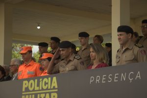 A imagem mostra diversas autoridades no palanque da solenidade de 55 anos da Afas. Na primeira fileira estão três homens e duas mulheres, sendo eles o chefe do Estado-Maior do CBMMG, coronel Moisés Magalhães de Sousa, e a comandante-geral do CBMMG, coronel Jordana de Oliveira Filgueiras Daldegan, ambos vestindo farda laranja; o comandante-geral da PMMG, coronel Frederico Otoni Garcia, que veste farda marrom e faz continência; a presidente da Afas, Mirella Cristina Ribeiro Garcia, com vestido vermelho e cabelos na altura dos ombros; e o chefe do Estado-Maior da PMMG, coronel Maurício José de Oliveira, também usando farda marrom.