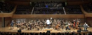 A imagem mostra a Sala Minas Gerais durante a apresentação da Orquestra Sinfônica da PMMG, com participação da banda 14 Bis e do coral Black to Black. Em cima do palco está a orquestra que conta com vários músicos sentados tocando diferentes instrumentos e vestindo farda marrom oficial da PMMG. A frente deles está o regente vestindo farda branca. Também estão os integrantes da banda 14 Bis nas extremidades do palco, tocando instrumentos. Na arquibancada superior está posicionado o coral Black to Black