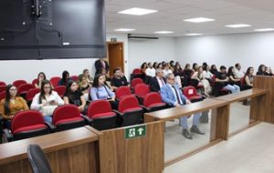 A imagem mostra os alunos do curso de Direito da Unifor-MG durante sessão no Tribunal Pleno. Eles estão sentados em cadeiras vermelhas. As paredes ao fundo são brancas e há uma bancada madeira em primeiro plano