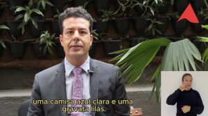 A imagem mostra o desembargador Fernando José Armando Ribeiro sentado, usando terno azul marinho, camisa clara e gravata lilás. Ao fundo há um jardim vertical com várias plantas. No canto direito inferior da imagem está a janela da intérprete de libras, de camisa preta e cabelos presos