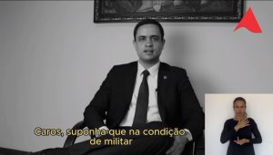 A imagem mostra o Juiz George Walter Barreto Paviotti sentado, de camisa branca, terno azul marinho e gravata verde. Ao fundo há uma parede branca com um quadro decorativo. No canto direito inferior da imagem está a janela da intérprete de libras, de camisa preta