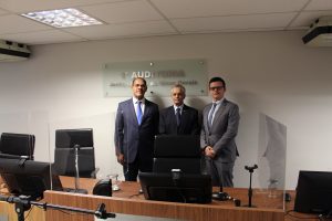 A imagem mostra três homens posando para foto na sala da Primeira Auditoria da Justiça Militar. O juiz Marcelo Adriano Menacho dos Anjos usa terno preto, gravata azul e óculos; o desembargador Sócrates Edgard dos Anjos veste terno preto com gravata azul escuro, e o juiz Bruno Cortez Torres Castelo Branco usa terno cinza com gravata da mesma cor e óculos. Eles estão atrás de cadeiras pretas e de uma tribuna de madeira com monitores de computador