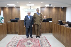 A imagem mostra o presidente do TJMMG, de terno cinza, e o comandante-geral da PMMG, de farda marrom da corporação. Ambos exibem no pescoço o Colar do Mérito Judiciário Militar. Eles estão posicionados no centro da sala do Tribunal Pleno, composta por tribuna e paredes de madeira, e um tapete estampado