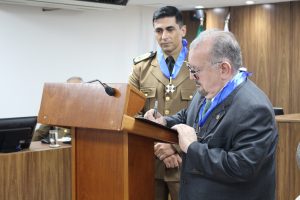 A imagem mostra o presidente do TJMMG e o comandante-geral da PMMG assinando um documento. O presidente usa terno cinza e está assinando um papel em um púlpito de madeira, sendo observado pelo comandante-geral, que veste uniforme marrom da corporação. Ambos exibem no pescoço o Colar do Mérito Judiciário Militar