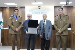 A imagem mostra o momento da entrega da placa em homenagem aos 250 anos da PMMG. Nela estão o comandante-geral e o chefe do Estado-Maior da PMMG vestindo farda marrom da corporação, tendo ao centro o presidente do TJMMG e o servidor que conduziu a placa, ambos vestindo terno cinza. O presidente do TJMMG segura nas mãos a placa de alumínio. Eles estão na sala do Tribunal Pleno do TJMMG, com tribuna e paredes de madeira