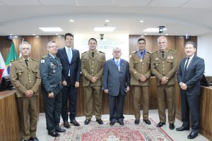 A imagem mostra um grupo de oito homens, entre os quais desembargadores do TJMMG civis e militares, além do comandante-geral e o chefe do Estado-Maior da PMMG. Eles estão na sala de audiências do Tribunal Pleno do TJMMG, em frente à uma tribuna e paredes de madeira