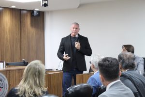 A imagem mostra o secretário de Estado de Justiça e Segurança Pública de Minas Gerais Rogério Greco discursando durante sua palestra. Ele fala ao microfone e veste camisa preta, blazer preto com calça jeans azul. Em primeiro plano estão os convidados da palestra; ao fundo há uma parede de madeira e uma parede branca
