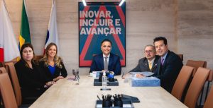 A imagem mostra cinco pessoas sentadas ao redor de uma mesa branca, sendo três homens e duas mulhes. Ao centro está o presidente da OAB-MG Gustavo Chalfun vestindo terno azul e gravata cinza. À direita estão os desembargadores do TJMMG, Jadir Silva usando terno preto, gravata amarela e óculos; e Fernando José Armando Ribeiro vestindo terno azul e gravata roxa. À esquerda está a ouvidora-geral da OAB-MG, Gláucia Fernandino usando sobretudo preto e a secretária-geral adjunta, Cássia Hatem vestindo blusa azul e blazer preto. Ao fundo há três bandeiras e um quadro colorido escrito "Inovar, incluir e avançar" dependurado na parede branca