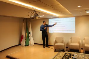 A imagem mostra o coronel Gilmar Luciano Santos ministrando aula no auditório do TJMMG. Ele usa camisa azul, calça preta, e fala ao microfone. Ele está apontando para o slide projetado na parede. À esquerda há três bandeiras e à direita há três poltronas brancas