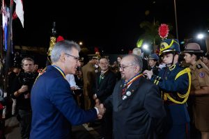 A imagem mostra o desembargador Jadir Silva vestindo terno preto com gravata listrada em preto e vermelho. Ele usa óculos e está recebendo a medalha Alferes Tiradentes de um oficial que veste farda. Ele cumprimenta o governador de Minas Gerais, Romeu Zema, que usa terno azul e óculos. Eles estão em um ambiente aberto e ao redor estão várias autoridades que também receberam a condecoração