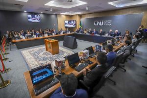 A imagem mostra a mesa de honra na sessão solene de comemoração aos 20 anos do CNJ, com uma ampla tribuna em formato retangular com computadores em cima. As autoridades estão sentadas em cadeiras pretas atrás da mesa e estão vestidas de maneira formal. Ao fundo há uma parede cinza, uma parede de madeira com televisões e um painel com o letreiro do Conselho Nacional de Justiça. Ao centro há um tapete colorido e um móvel de madeira