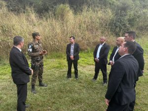 A imagem mostra seis militares que atuam no GSI do TJMMG recebendo capacitação de instrutor da 4ª Companhia de Polícia do Exército Brasileiro. Eles estão de pé em círculo em um campo aberto com grama e plantas ao fundo. Os militares vestem terno preto e o instrutor usa farda