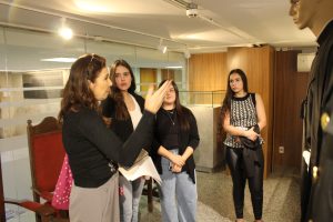 A imagem mostra quatro mulheres no Memorial do TJMMG, todas em pé, observando um manequim vestindo uma toga. Em primeiro plano está a servidora vinculada à Gestão Documental Maria Luiza Silveira Faria, que acompanhou o grupo. 