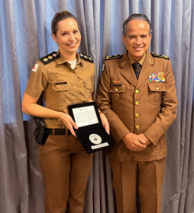 A imagem mostra a tenente-coronel Síria Delgado Matias e o desembargador James Ferreira Santos. Ambos vestem farda marrom. Ao fundo há uma cortina cinza.