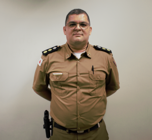 A imagem mostra o tenente-coronel Gustavo Costa Ferreira com as mãos cruzadas para trás. Ele usa farda marrom e óculos. A parede ao fundo é branca.