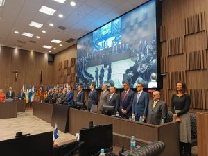 A imagem mostra 14 autoridades em destaque na abertura do III Encontro Interinstitucional do Eproc, em pé atrás de uma tribuna de madeira, sendo duas mulheres e 12 homens. O segundo da direita para a esquerda da imagem é o presidente do TJMMG, desembargador Jadir Silva, usando terno cinza, gravata amarela e óculos. Em primeiro plano há uma mesa com computadores e ao fundo há várias bandeiras. A parede é de madeira e possui um telão com a transmissão do evento