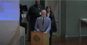 A imagem mostra o desembargador Rúbio Paulino Coelho falando em uma tribuna de madeira, ao microfone. Ele veste terno cinza e gravata vermelha