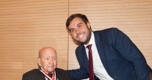 A imagem mostra o momento em que o juiz João Pedro Hoffert Monteiro de Lima entrega a Medalha do Mérito Judiciário Militar ao professor José Afonso da Silva. O homenageado está sentado, veste terno e gravata pretos. O juiz veste terno azul escuro com gravata vermelha. Eles estão atrás de uma mesa preta com uma placa com o nome do professor. A parede ao fundo é de madeira