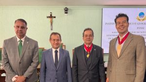 : Na imagem estão quatro homens posando para a foto em uma sala que, na parede ao fundo, há uma cruz e uma tela com informações do evento. Dois homens usam colares do IHGMG pendurados no pescoço. São eles o desembargador Fernando José Armando Ribeiro, que usa um blazer bege, e o desembargador José Marcos Vieira, de terno preto. Ao lado deles estão o desembargador Estevão Lucchesi, que usa terno cinza com gravata verde, e o presidente do TJMG Luiz Carlos Corrêa Junior, que usa terno azul marinho e blusa branca