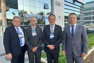 A imagem mostra quatro homens em frente ao edifício-sede do CNJ, em Brasília, um edifício com fachada de vidro e concreto cinza. O primeiro à esquerda da foto é o chefe de gabinete da Presidência, coronel Giovanne Gomes da Silva, vestindo terno preto e gravata azul. Ao seu lado está o corregedor da Justiça Militar mineira, desembargador Sócrates Edgard dos Anjos, que está com as mãos cruzadas à frente do corpo, e usa terno cinza com gravata listrada. O terceiro é o vice-presidente do TJMMG, desembargador James Ferreira Santos, que também está com as mãos cruzadas à frente do corpo. Ele veste terno preto e gravata cinza. No lado direito da imagem está o secretário especial da Presidência, Giovani Viana Mendes, vestindo terno cinza e gravata azul