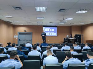A imagem mostra o juiz de Direito George Walter Barreto Paviotti em um palco, falando a uma plateia de aspirantes a oficiais da Aeronáutica. Ele está vestindo terno azul escuro com gravata azul claro, enquanto a plateia está sentada e veste uniforme azul. As paredes são marrons e ao fundo há um telão com a apresentação da disciplina