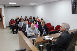 A imagem mostra o público presente na reunião, entre servidores e magistrados. Eles estão sentados em cadeiras de auditório vermelhas e assistem à apresentação na sala do Tribunal Pleno do TJMMG. Em primeiro plano está o desembargador Sócrates Edgard dos Anjos, corregedor, de terno.