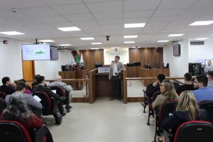 A imagem mostra servidor do TJMMG apresentando dados em telão durante reunião no plenário do TJMMG. Público acompanha atento. Ao fundo, à esquerda da imagem, está sentado o desembargador Sócrates Edgard dos Anjos. A sala é branca, com painéis e tribuna de madeira, e ao fundo há um painel com o nome do Tribunal por extenso