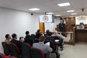A imagem mostra o corregedor de pé, vestindo terno, na sala do Tribunal Pleno do TJMMG. Ele fala aos magistrados que estão sentados em cadeiras vermelhas, atentos. A sala tem paredes brancas, painéis e tribuna de madeira e há duas telas exibindo o brasão da Justiça Militar de Minas Gerais.