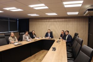 A imagem mostra seis pessoas, sendo quatro homens e duas mulheres, sentadas ao redor de uma mesa de reuniões na sala Diamante do TJMMG, com paredes revestidas de madeira clara e iluminação de teto. Os participantes, em trajes formais, estão atentos e voltados para a frente.
