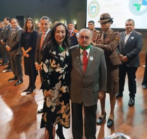 A imagem mostra um grupo de pessoas em pé, posicionado sobre um palco iluminado. Em destaque, à frente, estão a secretária de Estado de Meio Ambiente e Desenvolvimento Sustentável, de vestido preto com estampa floral branca e verde; e o presidente do TJMMG, que está de terno cinza com gravata vermelha, camisa verde e óculos. Ao fundo, diversos homens e mulheres, inclusive militares fardados, também posam solenemente.