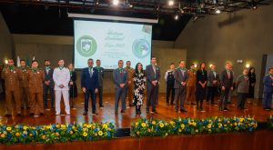 A imagem mostra o palco do Auditório JK com pessoas em pé e vestidas formalmente. Entre elas, há militares fardados e civis em trajes sociais, a maioria com medalhas penduradas no pescoço. Ao fundo, um telão exibe os dizeres “Destaque Ambiental – Edição 2025” e os logotipos do Governo de Minas Gerais e do Sistema Estadual de Meio Ambiente.