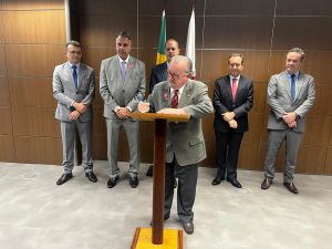 A imagem mostra o desembargador Jadir Silva assinando a portaria conjunta. Ele veste terno cinza e gravata listrada vermelha com preto. O palanque para a assinatura é de madeira e ao fundo há a bandeira do Brasil e de Minas Gerais. A parede é marrom e o chão é cinza