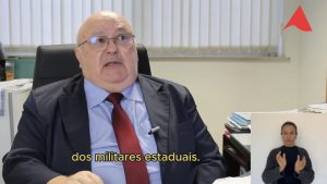 A imagem mostra o juiz João Libério da Cunha sentado, vestindo terno escuro e camisa clara. Ao fundo, há uma parede neutra com elementos institucionais. No canto inferior direito da imagem está a janela da intérprete de Libras, com camisa preta.