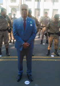 A imagem mostra o desembargador Rúbio Paulino Coelho, em ambiente externo, sob luz do dia, no pátio da Academia de Polícia Militar de frente para a câmera, com as mãos cruzadas à frente do corpo. Ele veste terno azul, camisa branca, gravata bordô e exibe a Medalha Cinquentenário EFO presa ao peito. Ao fundo, vários policiais militares uniformizados estão perfilados em frente a um prédio da APM, um prédio branco de fachada antiga, com janelas gradeadas e detalhes arquitetônicos verticais.