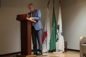 A imagem mostra o desembargador de terno cinza, de pé em um púlpito de madeira. Atrás há três bandeiras