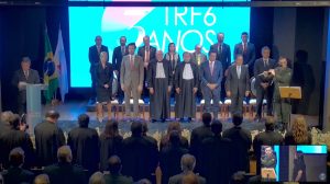 A imagem mostra o presidente do TJMMG, desembargador Jadir Silva, em pé, na primeira fileira de autoridades, sendo o antipenúltimo da esquerda para a direita. Ele veste terno cinza-claro e gravata amarela, participando da solenidade em comemoração aos 3 anos do TRF6. Ao seu lado, outras autoridades do Judiciário também estão posicionadas. Ao fundo, um painel eletrônico exibe a identidade visual do evento e, à esquerda, estão posicionadas as bandeiras do Brasil e de Minas Gerais. No canto direito da imagem, um músico da banda toca flauta durante a cerimônia