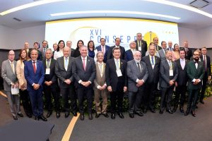 A foto mostra participantes do evento, entre homens e mulheres. Dentre eles está o vice-presidente do TJMMG, desembargador James Ferreira Santos, na primeira fila, vestindo terno azul e gravata vermelha. Ao fundo há uma parede branca com o letreiro escrito XVI Consepre