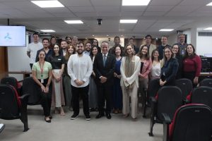 A imagem mostra um grupo de 25 pessoas posando para a foto na sala de audiências do TJMMG. Ao centro está o desembargador James Ferreira Santos, de terno preto, camisa branca e gravata azul com listras diagonais. Ao seu lado está o juiz João Pedro Hoffert Monteiro de Lima usando camisa branca e calça preta. Ao redor deles estão mulheres e homens, vestidos de forma social ou casual. Ao fundo, há uma parede de madeira e, à esquerda, uma tela exibe o logotipo da Rede Mineira de Inovação