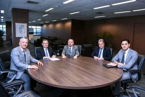 A imagem mostra cinco homens sentados ao redor de uma mesa redonda de madeira, em ambiente corporativo. Todos vestem terno e gravata. Da esquerda para a direita está o secretário especial da Presidência do TJMMG; o segundo é o presidente do TJMG, que segura uma caneta sobre um bloco de anotações; o terceiro é o presidente do TJMMG; o quarto é o chefe de gabinete da presidência do TJMMG; e o quinto é o diretor executivo de Finanças do TJMMG. Ao fundo, há paredes revestidas de madeira escura, poltronas pretas, um quadro com desenho em preto e branco e plantas decorativas. O teto é branco com luminárias embutidas