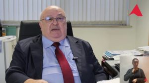A imagem mostra o juiz de Direito João Libério da Cunha, usando óculos e sentado em uma cadeira de escritório em seu gabinete. Ele veste terno azul-escuro, camisa azul-clara e gravata vermelha. Ao fundo, há uma persiana fechada e, à direita da imagem, papéis e pastas sobre uma mesa. No canto superior direito, há um detalhe da logomarca do TJMMG, destacando o triângulo da bandeira de Minas Gerais estilizado. No canto inferior direito, aparece um quadro menor com a intérprete de Libras de cabelo preso e usando camisa preta