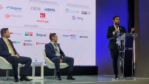 A imagem mostra a apresentação do projeto Banco de Sentenças. Mais à direita da imagem, em pé, atrás de um púlpito transparente e segurando um microfone, está o juiz João Pedro Hoffert Monteiro de Lima durante sua palestra. Ele veste terno preto, camisa branca e gravata azul-clara. À esquerda estão dois homens de terno, sentados em poltronas brancas. Ao fundo, há um painel luminoso com diversas logomarcas.