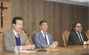 A imagem mostra três homens sentados em uma mesa de madeira. O primeiro à esquerda é o desembargador Fernando José Armando Ribeiro, que fala ao microfone. Ele veste paletó de lã cinza com suéter azul por baixo. Ao centro está o juiz Richardson Xavier Brant, que veste terno azul-claro, camisa branca e gravata azul. À direita, o professor Marcelo Albuquerque, que usa terno preto com camisa branca e gravata preta com bolinhas