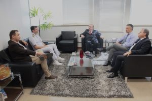 A imagem mostra cinco homens sentados em poltronas pretas na sala da Presidência do TJMMG. Eles estão dispostos ao redor de uma mesa de centro de vidro, sobre um tapete cinza. Ao centro está o presidente do TJMMG, que veste terno preto com gravata vermelha. À esquerda da imagem está o assessor de Relações Institucionais da Capitania dos Portos, de terno marrom, camisa clara e calça creme, e o capitão de mar e guerra, também da Capitania dos Portos, com uniforme branco da Marinha. Do lado direito estão o chefe de gabinete da Presidência do TJMMG, de terno preto e camisa branca, e o secretário especial da Presidência, de camisa social, calça e gravata cinza. Ao fundo, a sala é iluminada por janelas grandes com persianas brancas.