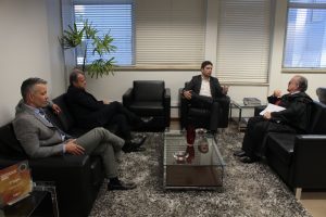 A imagem mostra quatro homens sentados em poltronas pretas na sala da Presidência do TJMMG. Eles estão dispostos ao redor de uma mesa de centro de vidro, sobre um tapete cinza. Ao centro está o controlador-geral do Estado, que veste bleiser e calça preta, com camisa clara. À direita da imagem está o presidente do TJMMG, que veste toga preta com gravata vermelha, e segura papéis. À esquerda da imagem está o chefe de gabinete da Presidência do TJMMG, de terno preto e camisa branca, e o secretário especial da Presidência, de camisa social, calça e gravata cinza. Ao fundo, a sala é iluminada por janelas grandes com persianas brancas.