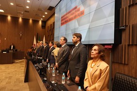 A imagem mostra autoridades em destaque alinhadas em pé no auditório do TJMG, durante a solenidade de abertura do Encontro de Juízes e Juízas Criminais e de Execução Penal. O juiz de Direito da Justiça Militar de Minas Gerais Marcos Luiz Nery Filho, que aparece em segundo lugar da direita para a esquerda, veste terno cinza escuro, camisa clara e gravata. Atrás das pessoas está um grande painel eletrônico com a identidade visual do evento