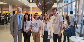 A imagem mostra cinco pessoas, entre homens e mulheres, integrantes do Comitê PopRuaJud durante a ação “Rua de Direitos”. Ao centro está a juíza Carolina Aleixo Benetti de Oliveira Rodrigues de óculos, vestindo calça bege e camiseta branca do evento. Os outros integrantes também usam a blusa do evento, exceto um homem que veste terno bege com camisa azul-clara e gravata listrada. Eles estão no hall de um edifício com grandes janelas de vidro. Ao fundo, participantes circulam pelo espaço e ao centro do hall se destaca uma escultura metálica